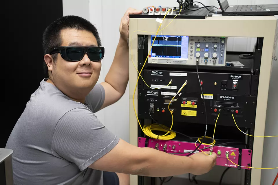 Yicheng Shi wears dark safety glasses as he poses next to a rack of electronics equipment.