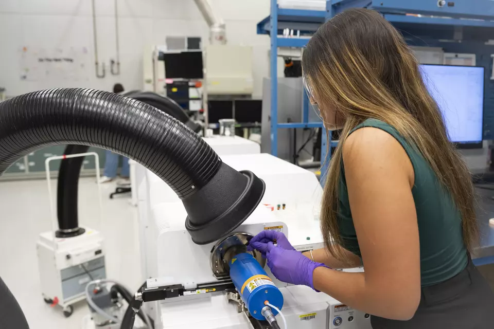 A female researcher with long hair stands working at a large piece of laboratory equipment, holding a small vial of material to be analyzed.