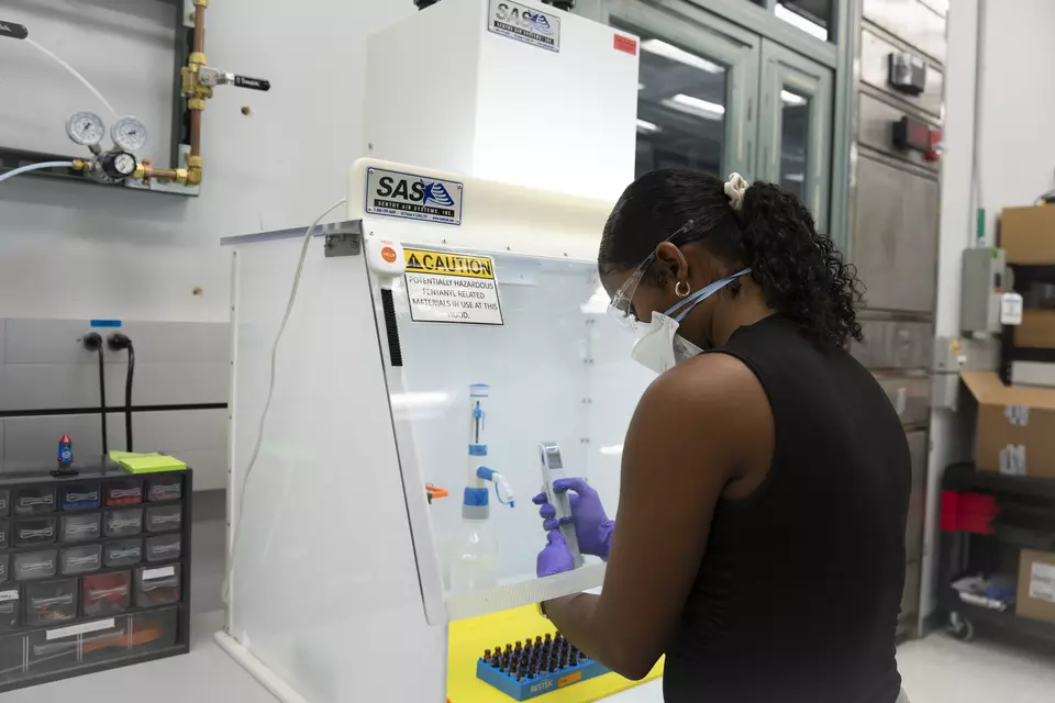 A researcher wearing safety glasses and a face mask works with her hands inside a plastic box, using a device to extract material from small vials.