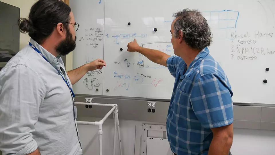Two men face away from the camera, pointing to diagrams written on a whiteboard. 