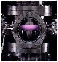 Photograph of a vacuum chamber with bolted flange window and violet plasma irradiating a surface.