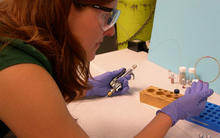 Woman with safety glasses and purple gloves working in lab. In her left hand she has a device with a tube attached to it. The tube is running into a small vial. In her right hand she holds a coiled tube leading from the vial.