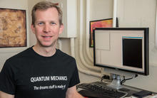 Scott Glancy, a sandy blonde-haired Caucasian man with blue eyes, standing in his office wearing a black T-shirt that reads "Quantum physics: The Dreams Stuff is Made of