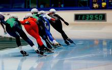 Five speed skaters making a turn on the track. A clock reading "11:39:50" is visible on the wall in front of them. 