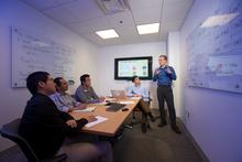 man standing in front of a white board filled with equations. Four other men sit around a table watching.