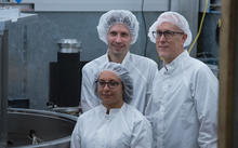 two men and a woman inside a lab and wearing protective white suits and hair nets look toward something out of frame