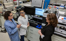 Carolyn Burdette, Laura Regalado and Katrice Lippa having an animated discussion in the lab. Behind them is a computer screen and other laboratory equipment, including a liquid chromatography mass spectrometer. 