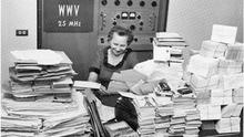 a woman sitting in front of a control panel that reads "WWV 2.5 MHz." In front of her is a table covered with hundreds of letters
