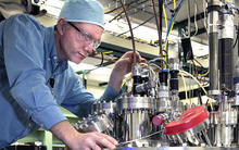 man with goggles, blue shirt and blue cap adjusts a pipe on top of a gleaming aluminum ultra-high vacuum (UHV) metal deposition chamber