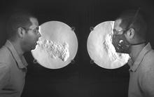 two photos of Matt Staymates facing each other in front of a schlieren camera system. On the left, Matt is not wearing a mask, on the right he is. In both photos he is coughing. In the unmasked photo, you can see a big plume of air exiting forward like a jet from his mouth. In the masked photo, the air plume hangs around his face and does not go very far. 