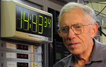 NIST physicist Judah Levine with the NIST time scale, a digital clock face reading 14:33:34 and a bank of atomic clocks. 