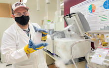 masked man in a white lab coat holding up a frozen container in a laboratory