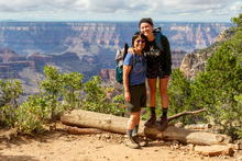 Two women in hiking gear stands at the top of a canyon, which is in the background.