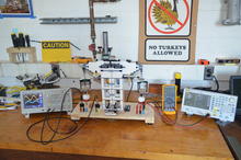 A Lego structure wired to various electronic readout devices stands on a wooden counter surrounded by other scientific equipment. A sign on the concrete wall behind reads "No Turkeys Allowed."