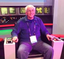 Barry Schneider poses sitting in a command chair on a replica of the bridge of the Starship Enterprise. 