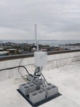 On a rooftop overlooking a waterfront town, an equipment box is attached to a pole anchored with concrete blocks. 