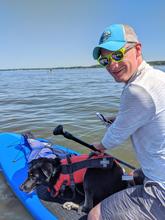 Ed Sisco smiles while floating on a paddleboard with a dog in a life vest. 