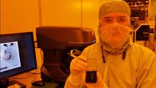 Under the orange lights of a clean room, Katarina Cicak, wearing a coverall and safety glasses, displays tiny chips in a plastic box.