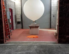 A large white balloon with a small box attached to it waits inside a cinderblock building with a rolltop door. 