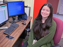 Isabelle Rivera poses smiling at a computer with two monitors, one showing a spreadsheet and the other multiple file icons. 