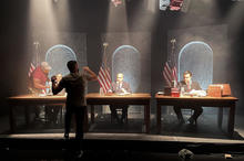 A man faces away from the camera, gesturing as he speaks to three actors seated behind desks on a film set. 