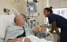 A smiling, female health care worker tends to an elderly male patient in a hospital bed.