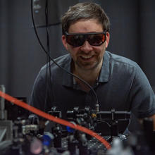 Krister Shalm, wearing safety glasses, smiles over a lab table covered with electronics equipment.