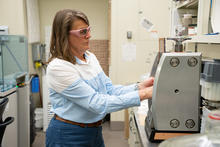 A researcher wearing safety glasses stands in the lab, reaching into a metal device on the lab table. 
