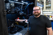 A bearded man poses smiling next to an open cabinet of equipment in a lab. 