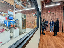 Four people stand talking in a converted factory space near a large interior window through which a complex, high-tech manufacturing line is visible. 