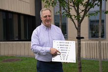 Jack Glover poses outside on the NIST campus, holding a flat square object with holes of varying sizes. 