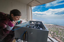A researcher peers into a box of scientific equipment near an opening in the wall showing the view down the mountain. 