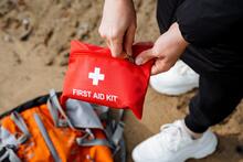 A person holds a red pouch labeled First Aid Kit as they stand over a travel bag on sandy ground.