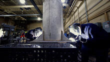 Two people wearing welding masks and other safety equipment bend over a table to work on a large metal cylinder.