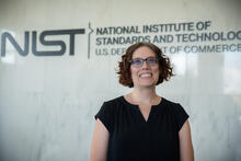 A woman with short curly brown hair and glasses smiles at the camera. NIST logo is in the background. 
