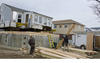 Men working at construction site with wood planks on ground and gray house dangling in air by crane