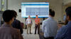 Two men stand in front of an audience, demonstrating WIPP, which is projected via computer onto several large screens in a library.