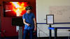 Person wearing a VR headset and holding controllers. Behind him a screen shows what the researcher is seeing within the headset--a virtual office building that is on fire.