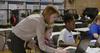 woman stands over the shoulder of two students working on laptops. 3D printers in the background.