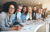 Creating Value for ALL Key Stakeholders Photo of a group of diverse employees behind a table standing together in a row in a bright office.