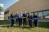 Six people stand in front of a tan building, cutting a blue ribbon