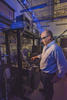 A man in safety glasses stands near a rack of equipment in a darkened lab.