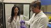 A woman in a lab coat (left) watches as a man in a lab coat (right) gestures at a small device he is holding in his hand.  