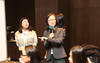 Hae-Jeong Lee speaks into a microphone as a student stands beside her