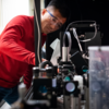 A man (Young Jong Lee) in a red shirt and safety goggles leans over a piece of scientific equipment.