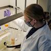 A woman (Alix Rodowa) wearing a mask and a lab coat works with scientific equipment.