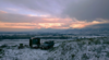 A small pickup truck and trailer stand on a grassy slope with snow and mountains in the background.