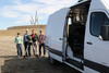 Four people stand around an instrument on a tripod next to the open door of a white van.