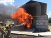 Firefighters spray water into a burning concrete building with a sign saying "NIST Fire Research."