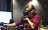 woman in a plaid shirt using a set of VR goggles and hand controls, a man sits at a computer in the background